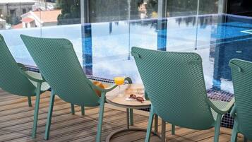 A glass-enclosed pool area with green chairs and a small table with a drink.