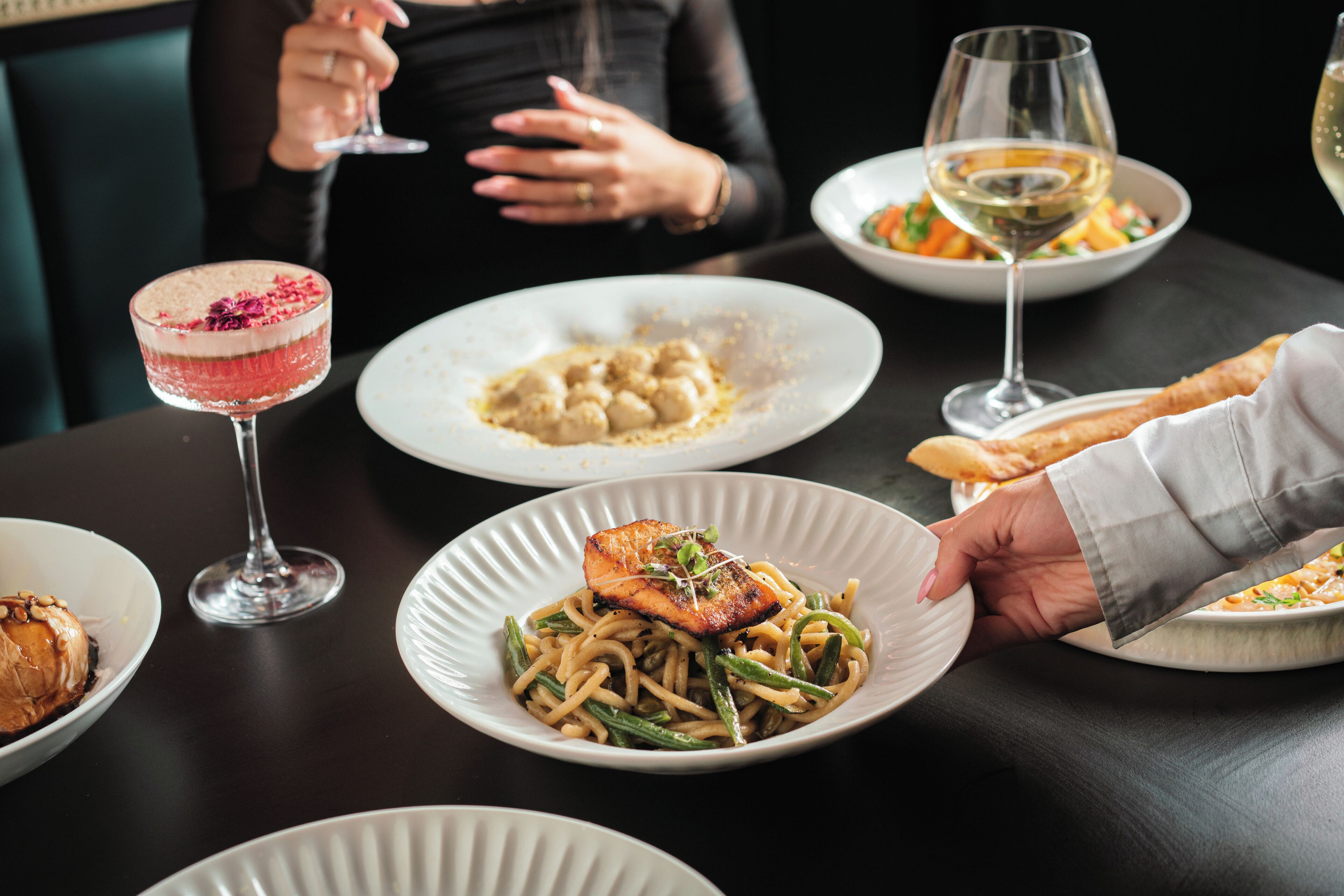 Una mesa de comedor con diversos platos, una copa de vino y una persona alcanzando un plato.
