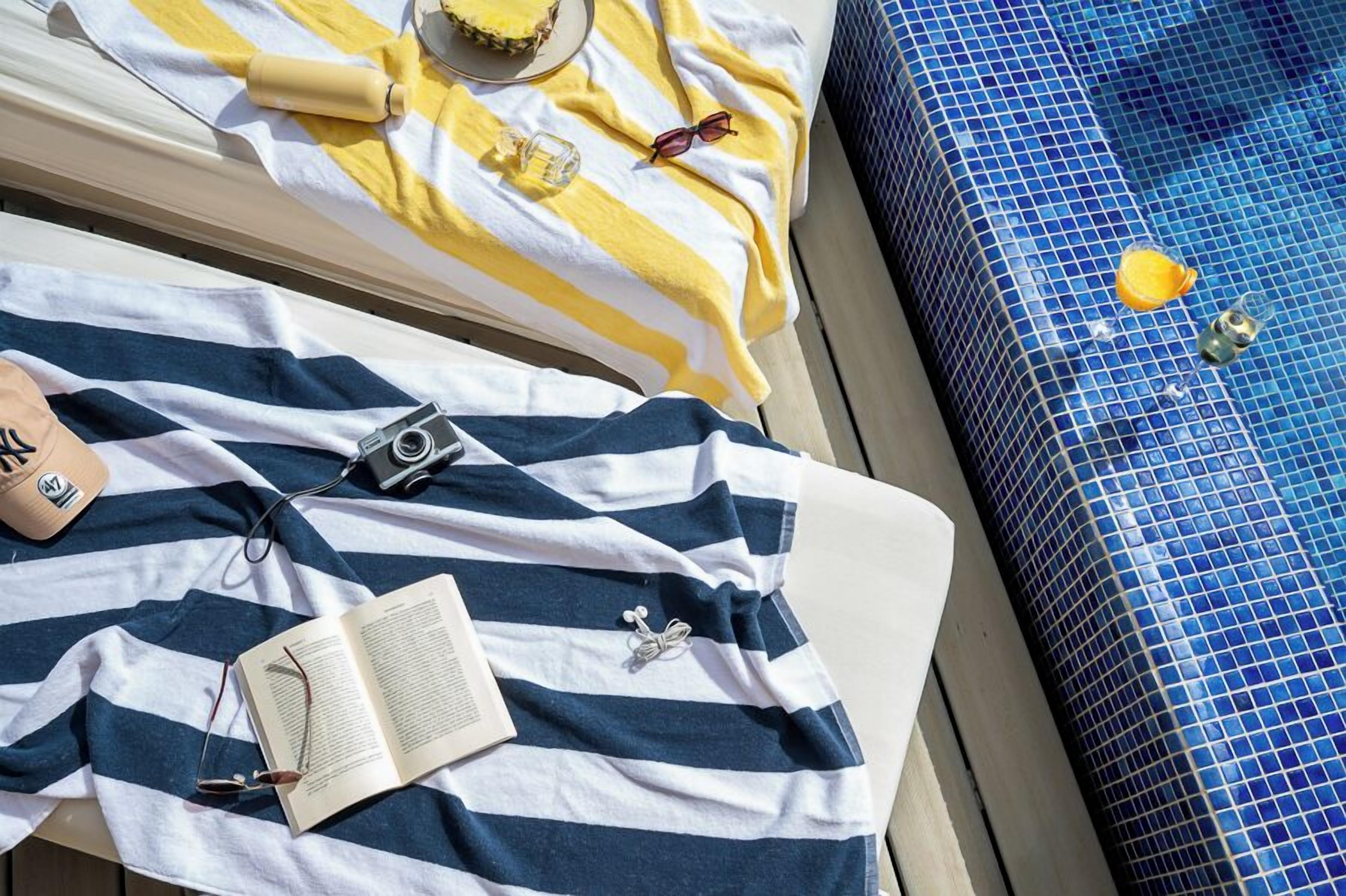 Una tumbona con toallas a rayas, un libro, gafas de sol, una cámara y una botella de protector solar.