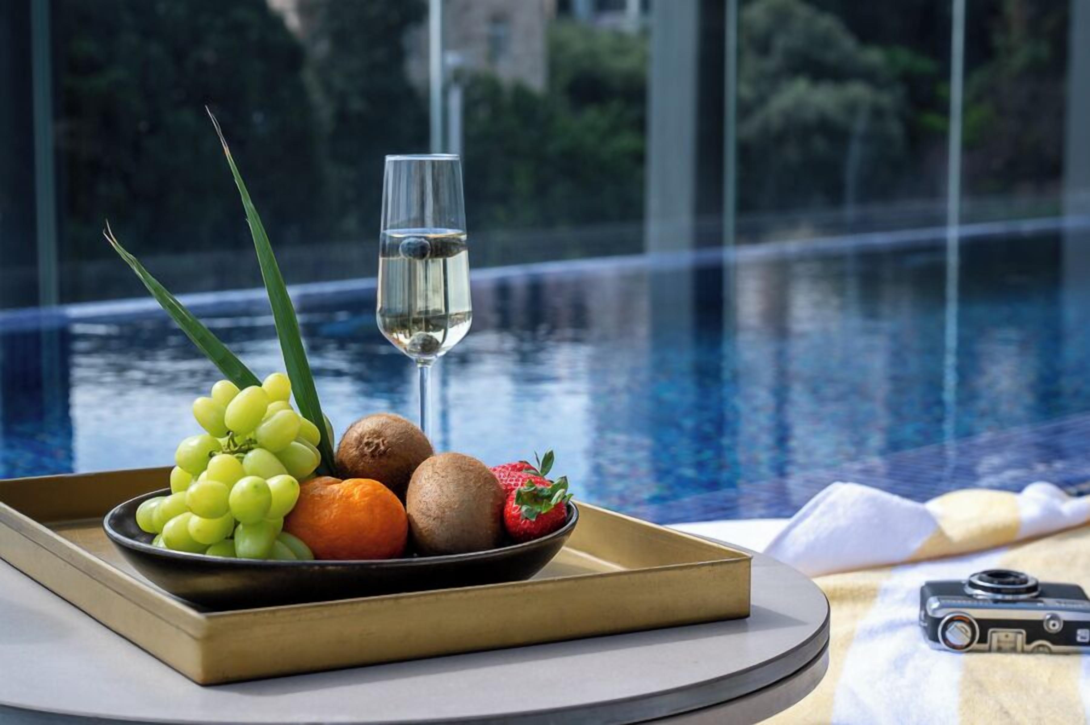 A tray with fruits and a glass of white wine by a poolside with a camera on the table.