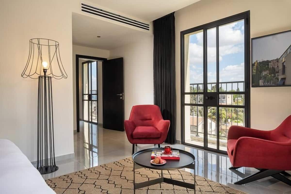 Una habitación de hotel moderna con un ventanal grande, un sillón rojo, una lámpara de pie negra y una mesita con un libro y un plato.