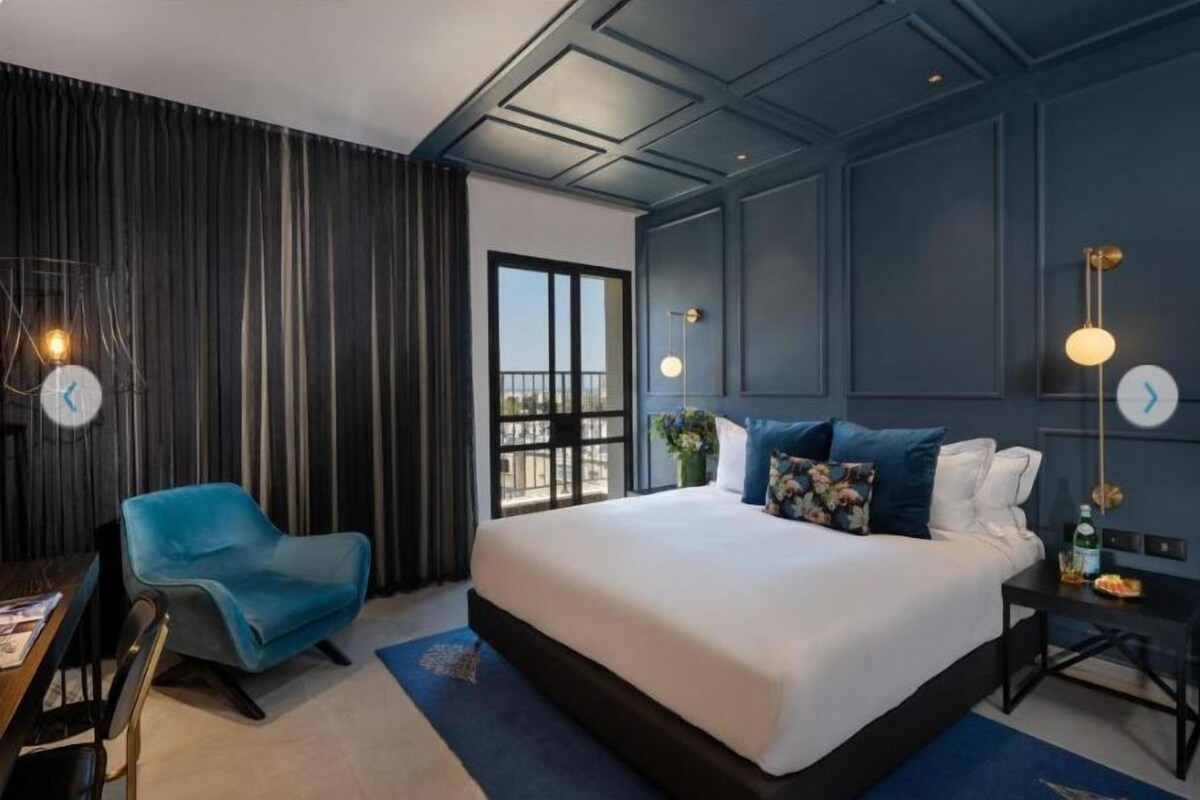 A modern hotel room with a large bed, a blue armchair, a desk, and a view of the city through a large window.