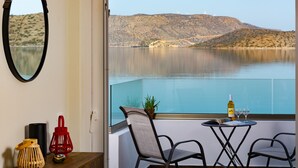 Un balcone con vista su un lago e delle colline, con due sedie, un tavolo con una bottiglia e dei bicchieri, e alcuni oggetti decorativi.