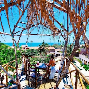 A person sitting at a wooden table under a thatched roof, overlooking a coastal area with buildings and palm trees.