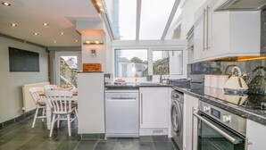 A modern kitchen with a dishwasher, washing machine, and oven. There is a dining area with a table and chairs. - 6 Milton Place (Bideford)