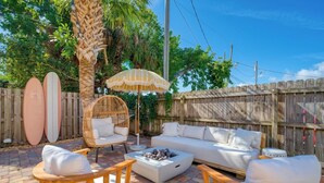 A patio area with a wicker chair, a white sofa, and a round table. There are two surfboards leaning against a wooden fence. - Luxury Villa, 2.5 Blocks from the Beach, Hot Tub, Outdoor Oasis, Mini Putt, Arcade games and so much more. (Cape Canaveral)