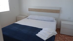 Una cama bien hecha con una manta azul y almohadas blancas, un cabecero de madera y dos mesitas de noche con cajones blancos.