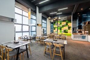 Un comedor moderno con mesas, sillas, ventanas amplias y una pared verde con plantas.