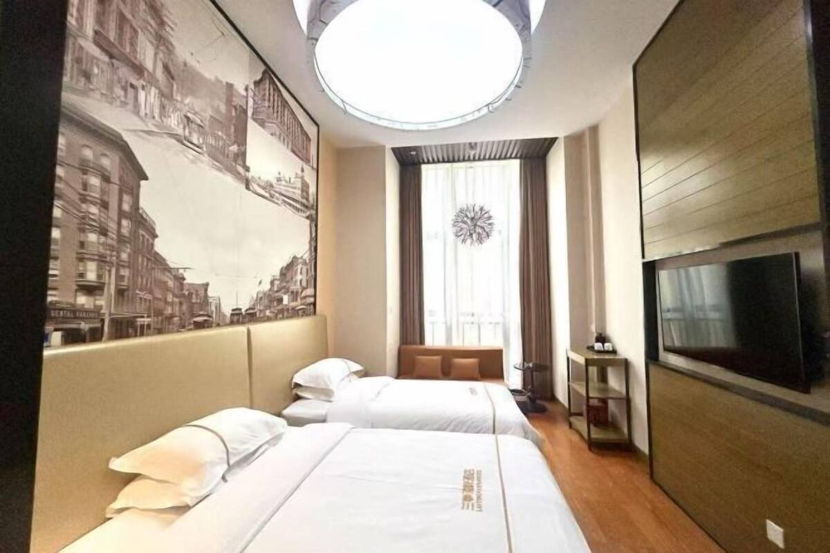 Quarto de hotel com duas camas, televisão, mesinha e uma janela grande com cortinas.