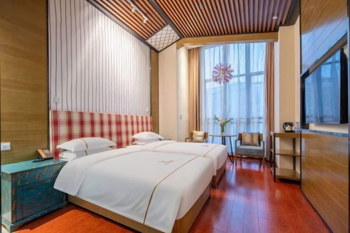 Quarto de hotel com duas camas, teto de madeira, janela com cortinas, uma mesa pequena e uma televisão.