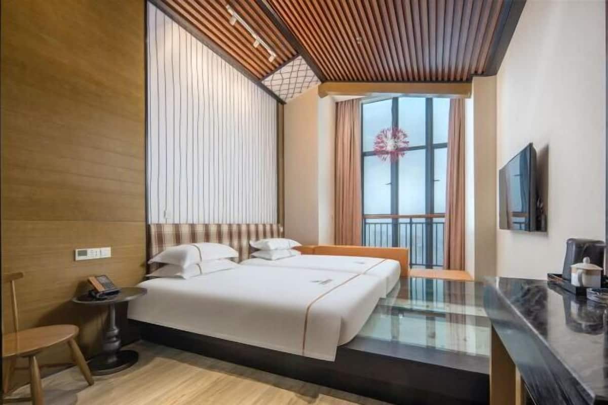Quarto de hotel com cama, escrivaninha, cadeira, janela com cortinas e televisão na parede.