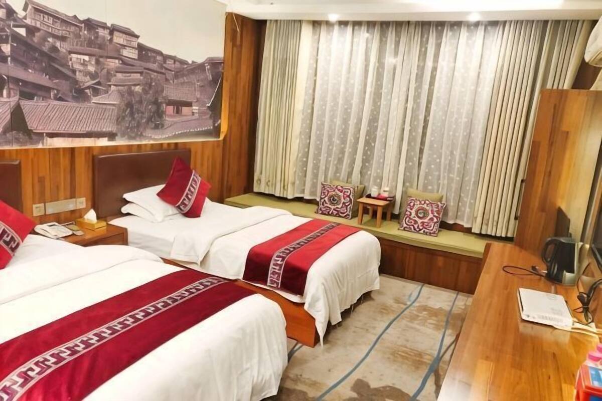 A hotel room with two beds, a wooden desk, a mirror, a window with curtains, and a wall-mounted TV.