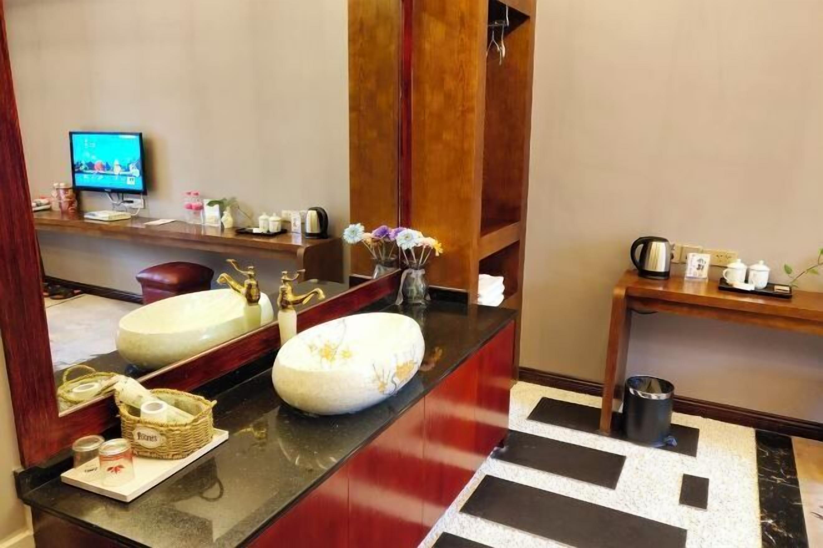 A hotel bathroom with a large mirror, two sinks, a television, and a wooden vanity with a vase of flowers.