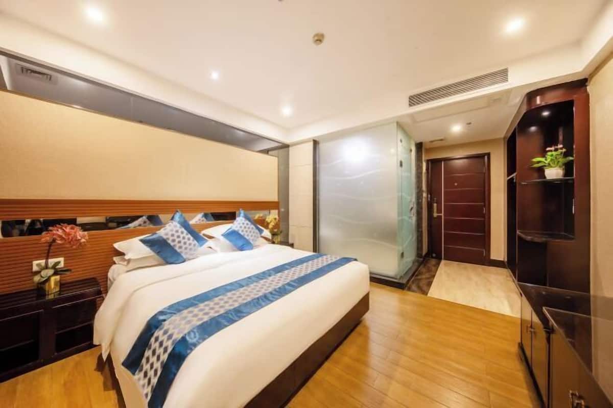 A hotel room with a large bed, wooden floor, a nightstand with a vase, a wardrobe, and a small plant on a shelf.