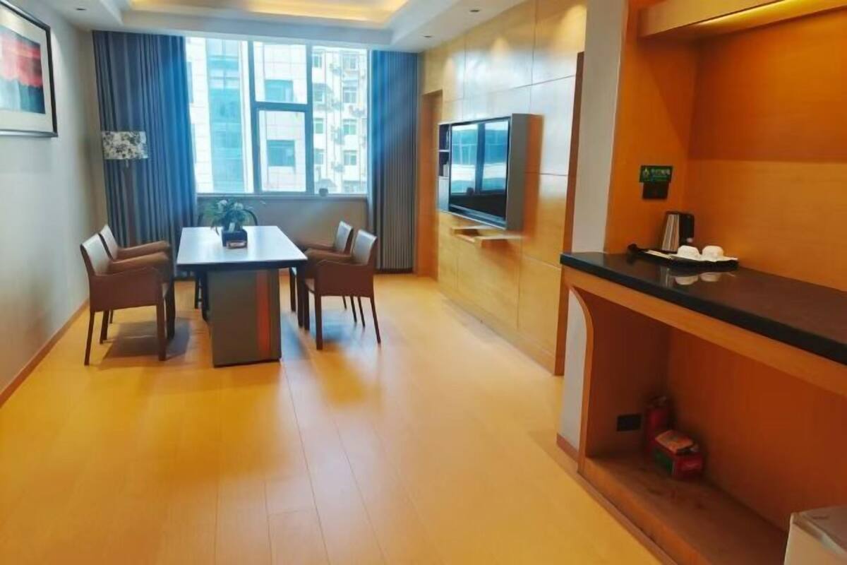 A modern hotel room with a dining area, a flat-screen TV, a minibar, and a large window with curtains.