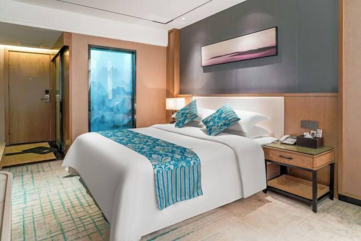 A modern hotel room with a large bed, a bedside table with a lamp, and a cityscape view through a glass door.