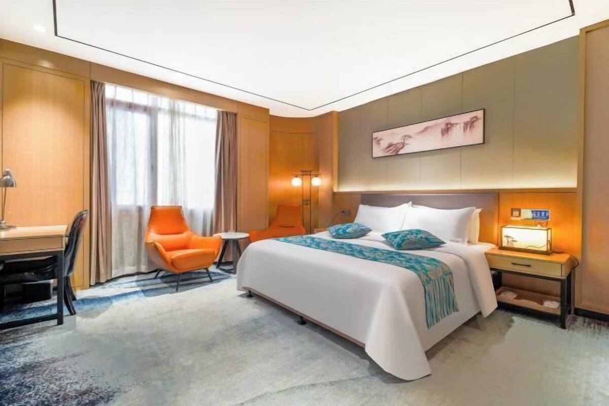 A hotel room with a bed, a desk, an orange chair, and a bedside table with a lamp.