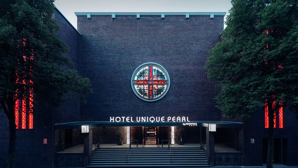 La entrada del HOTEL UNIQUE PEARL by ADOMOTELS, con un logotipo circular destacado sobre la puerta y fachada de ladrillo.