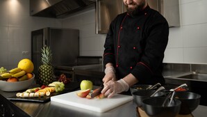 Un chef en una cocina profesional con electrodomésticos de acero inoxidable, una tabla de cortar blanca con fruta y diversos utensilios de cocina.