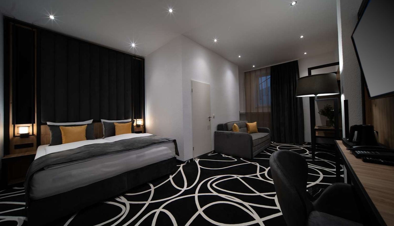 A modern hotel room with a large bed, a desk, a chair, and a sofa.