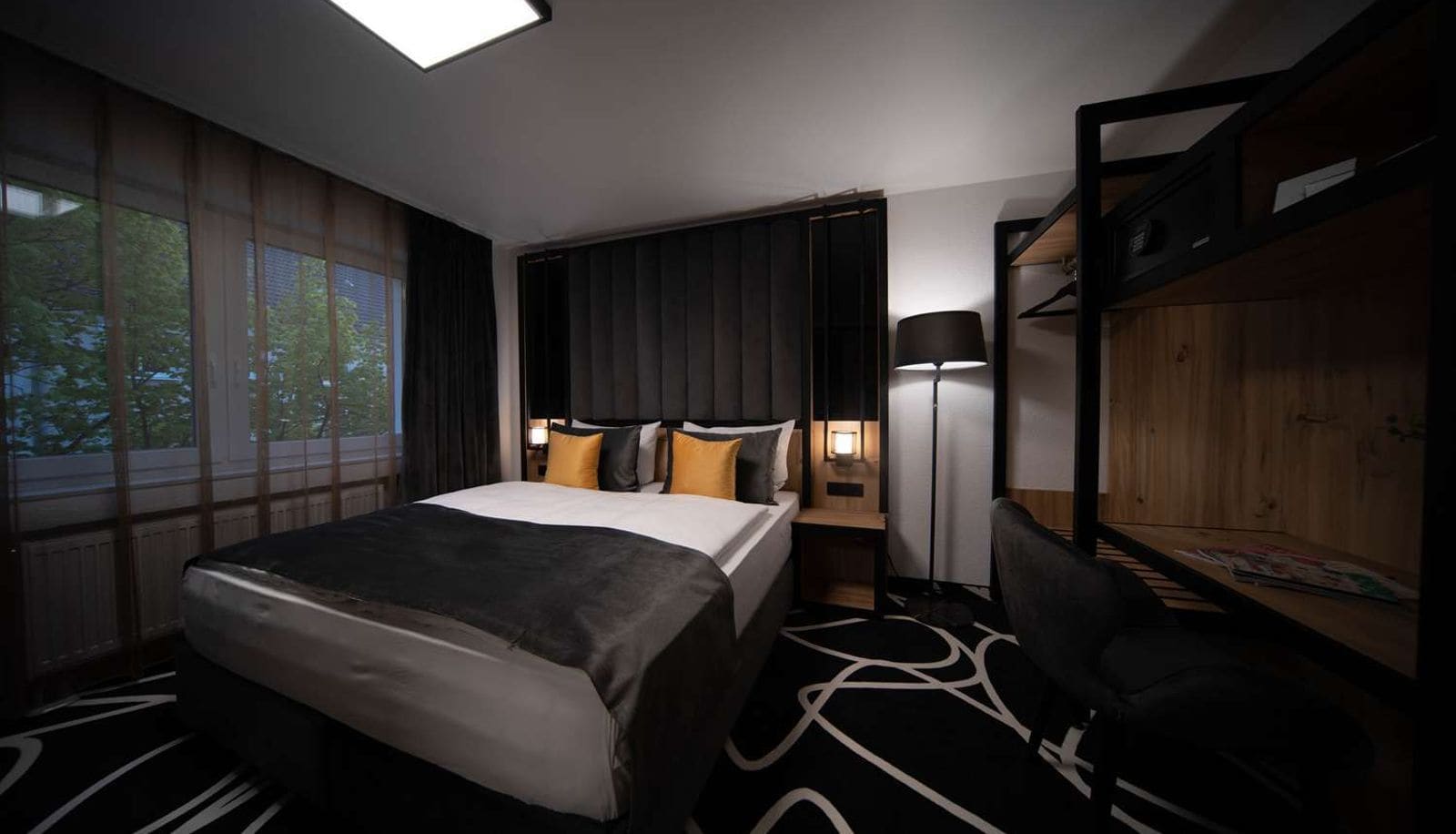 A hotel room with a bed, bedside table, lamp, and a view of the outdoors.