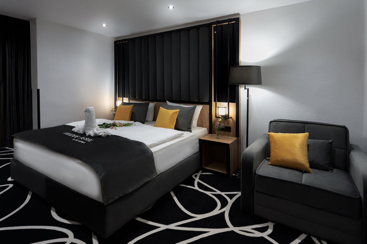 Une chambre d’hôtel moderne avec un grand lit, une table de chevet avec une lampe, un canapé gris et une moquette à motifs.
