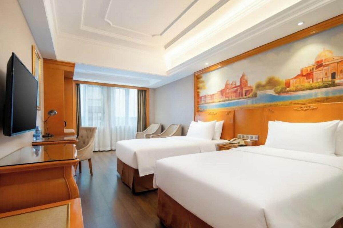 A hotel room with two beds, a desk, a TV, and a painting of a building on the wall.