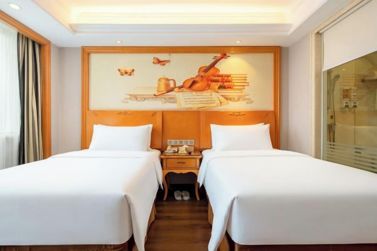 A hotel room with two beds, a wooden headboard, a nightstand with a lamp, and a window with curtains.