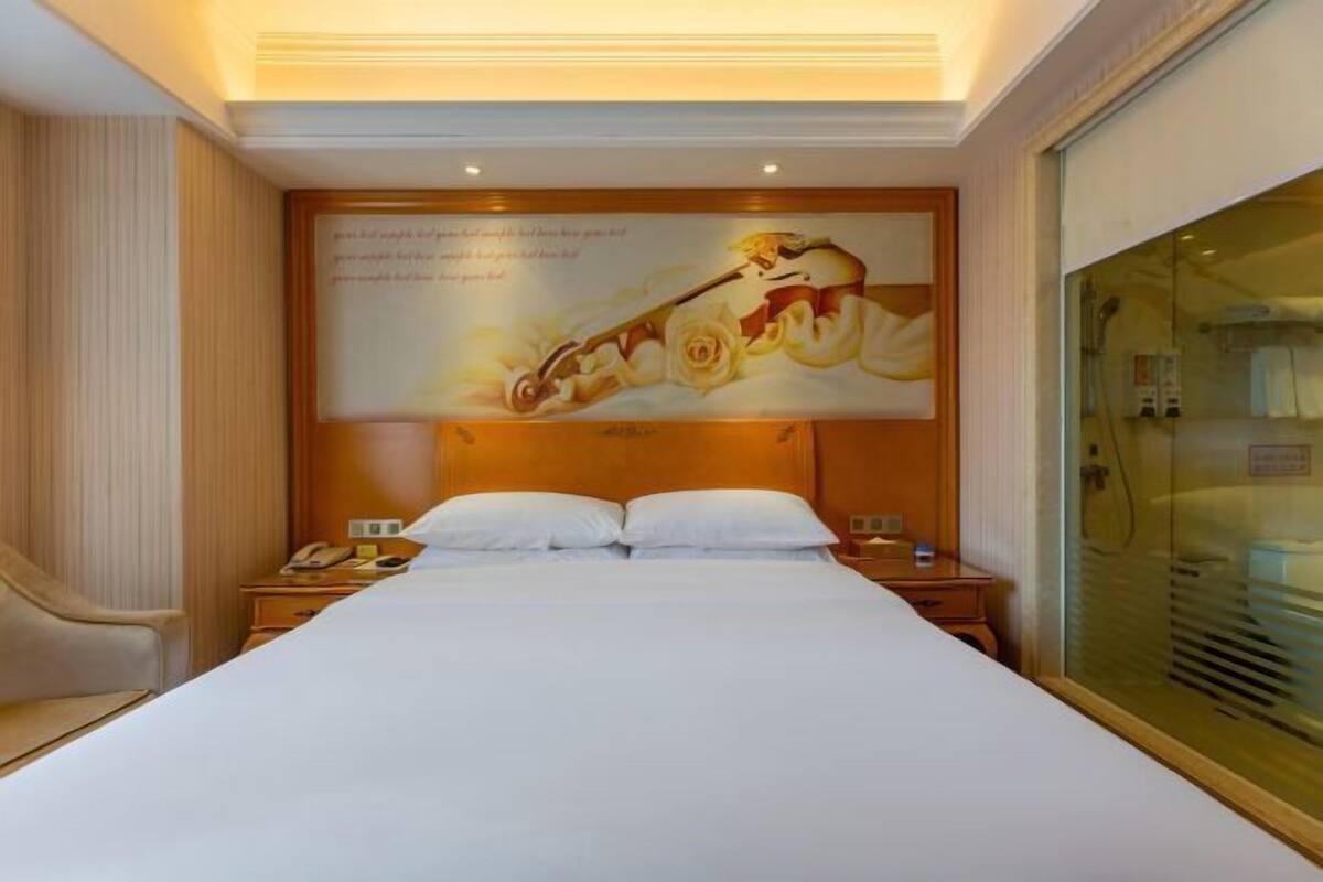 Une chambre d’hôtel avec un grand lit, une tête de lit ornée d’une fresque, une table de chevet avec un téléphone et une cloison en verre.