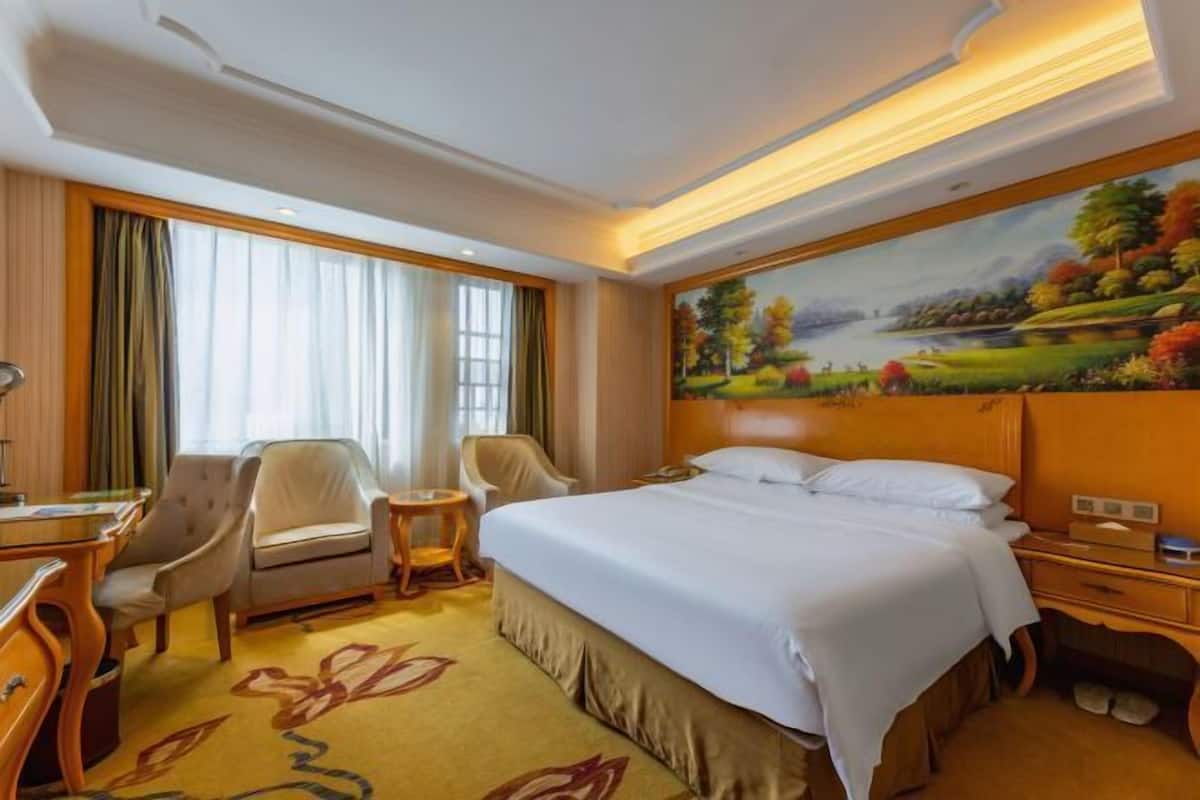 Une chambre d’hôtel avec un grand lit, un bureau, deux chaises, une petite table et un tableau représentant un paysage accroché au mur.