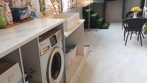Zona de lavandería con lavadora, estantes con productos de limpieza y comedor con sillas y mesa.
