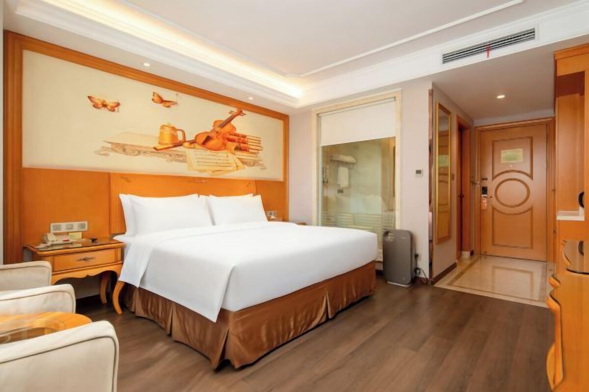 A hotel room with a large bed, a nightstand, a wall-mounted artwork, and a door leading to another room.