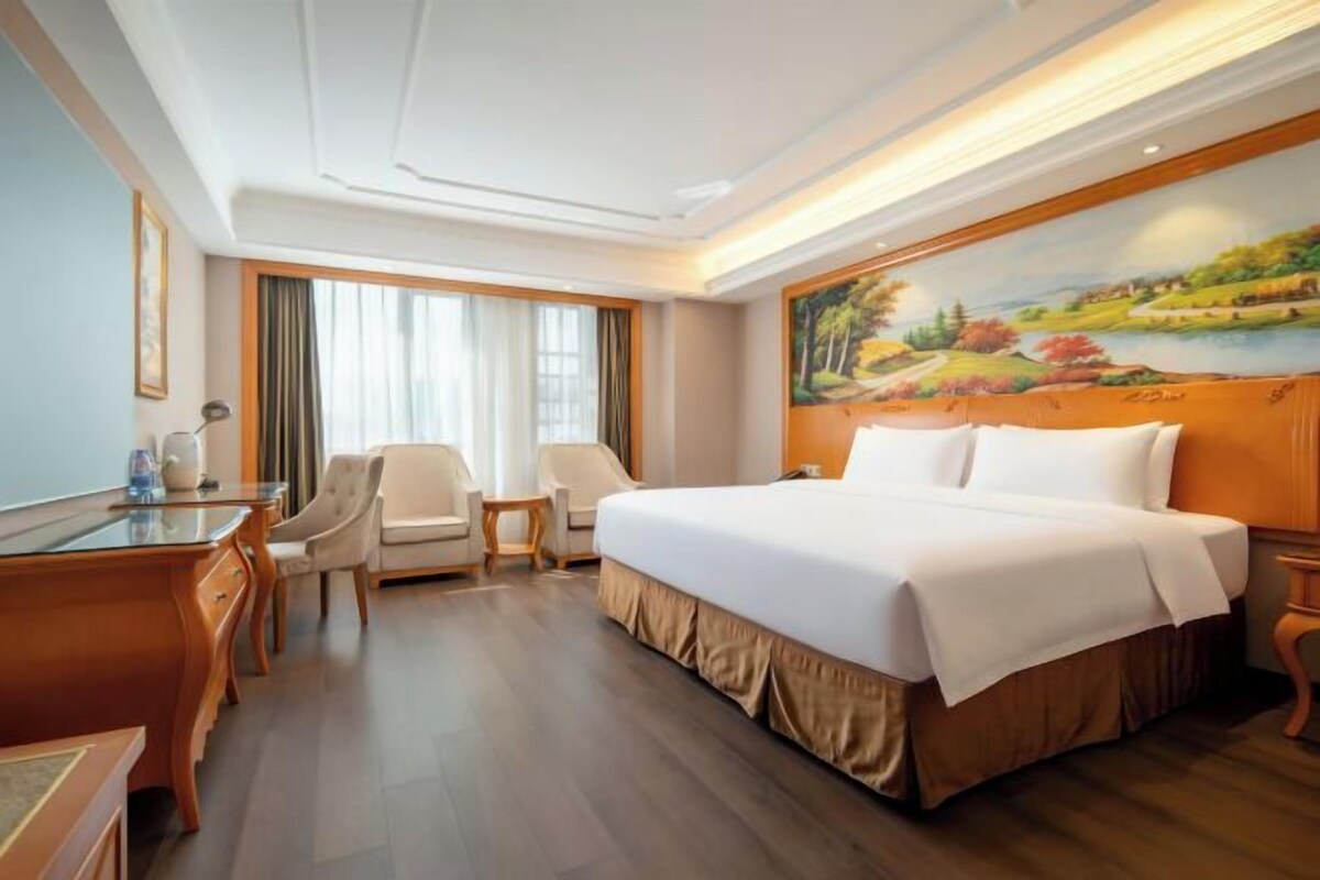 A hotel room with a large bed, two chairs, a desk, and a painting on the wall.