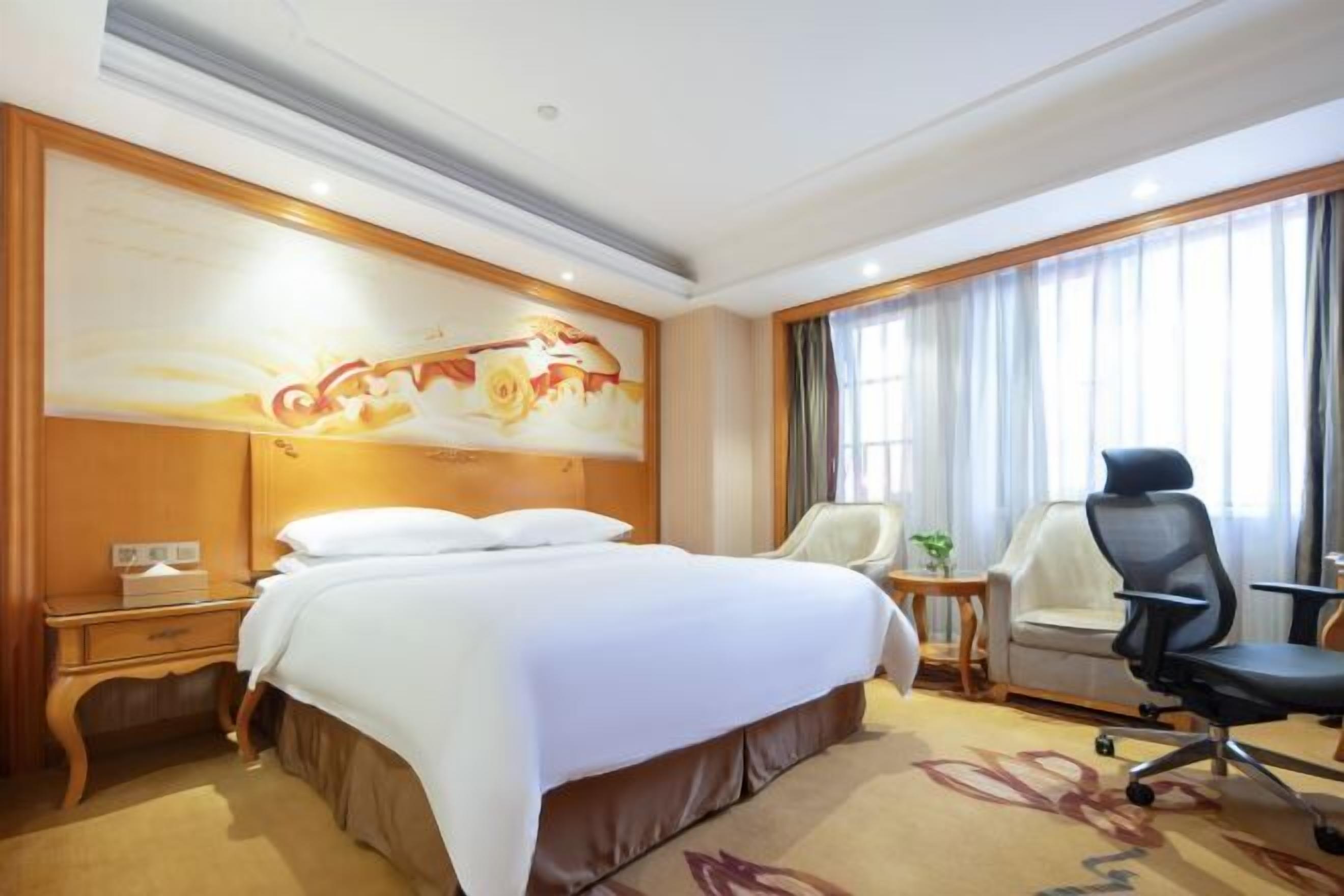 A hotel room with a large bed, a desk, a chair, and a painting on the wall.