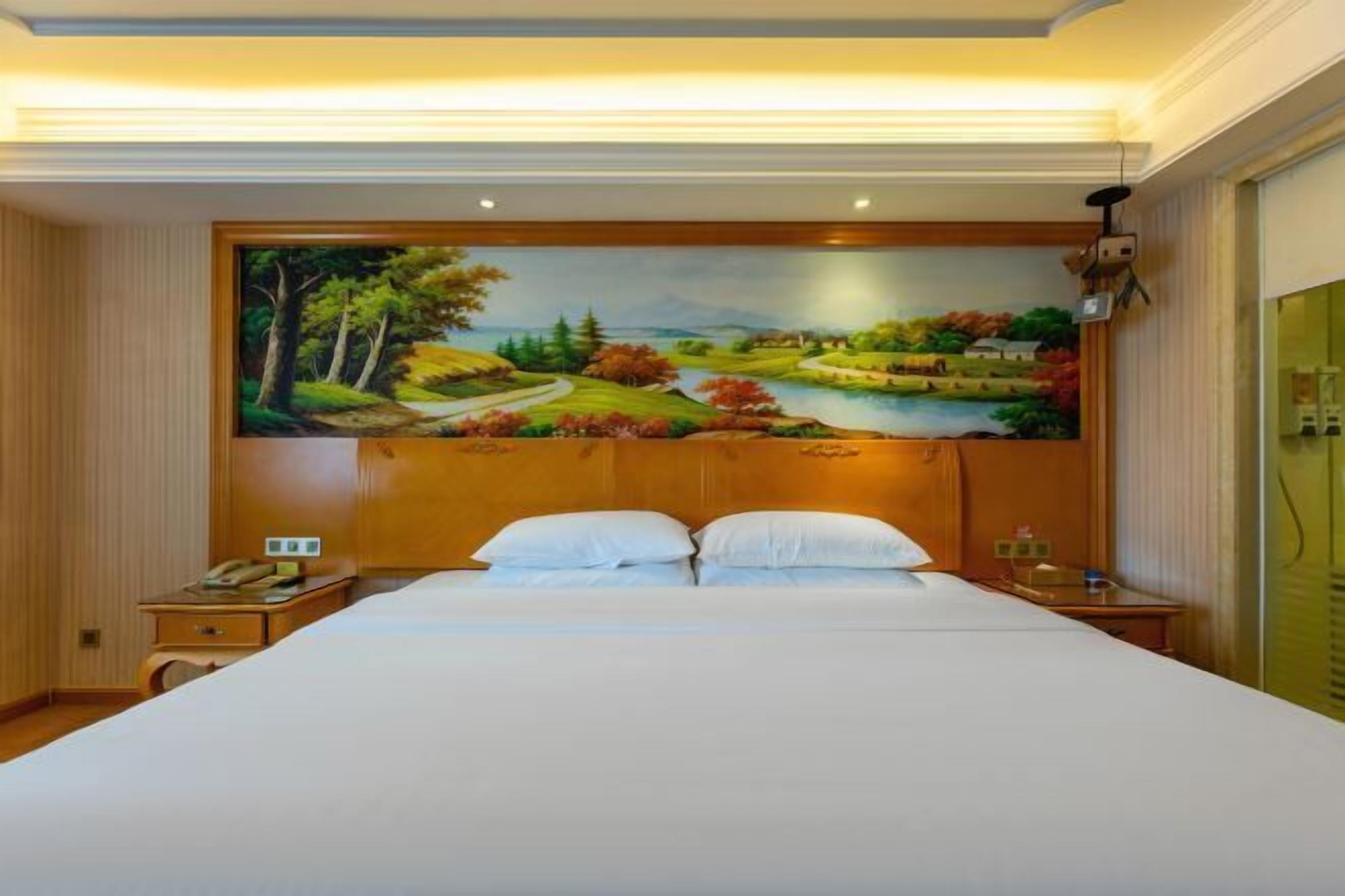 A hotel room with a large bed, two bedside tables, a painting of a landscape on the wall, and a wooden headboard.