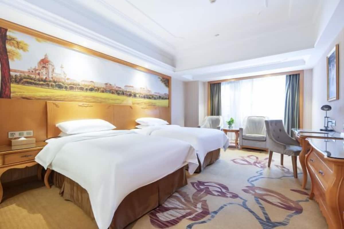 Une chambre d’hôtel avec deux lits, un bureau, une chaise et un grand tableau au mur.