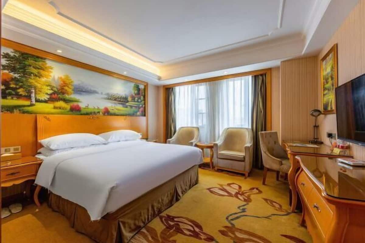 Une chambre d’hôtel avec un grand lit, deux fauteuils, un bureau, une télévision et un tableau au mur.