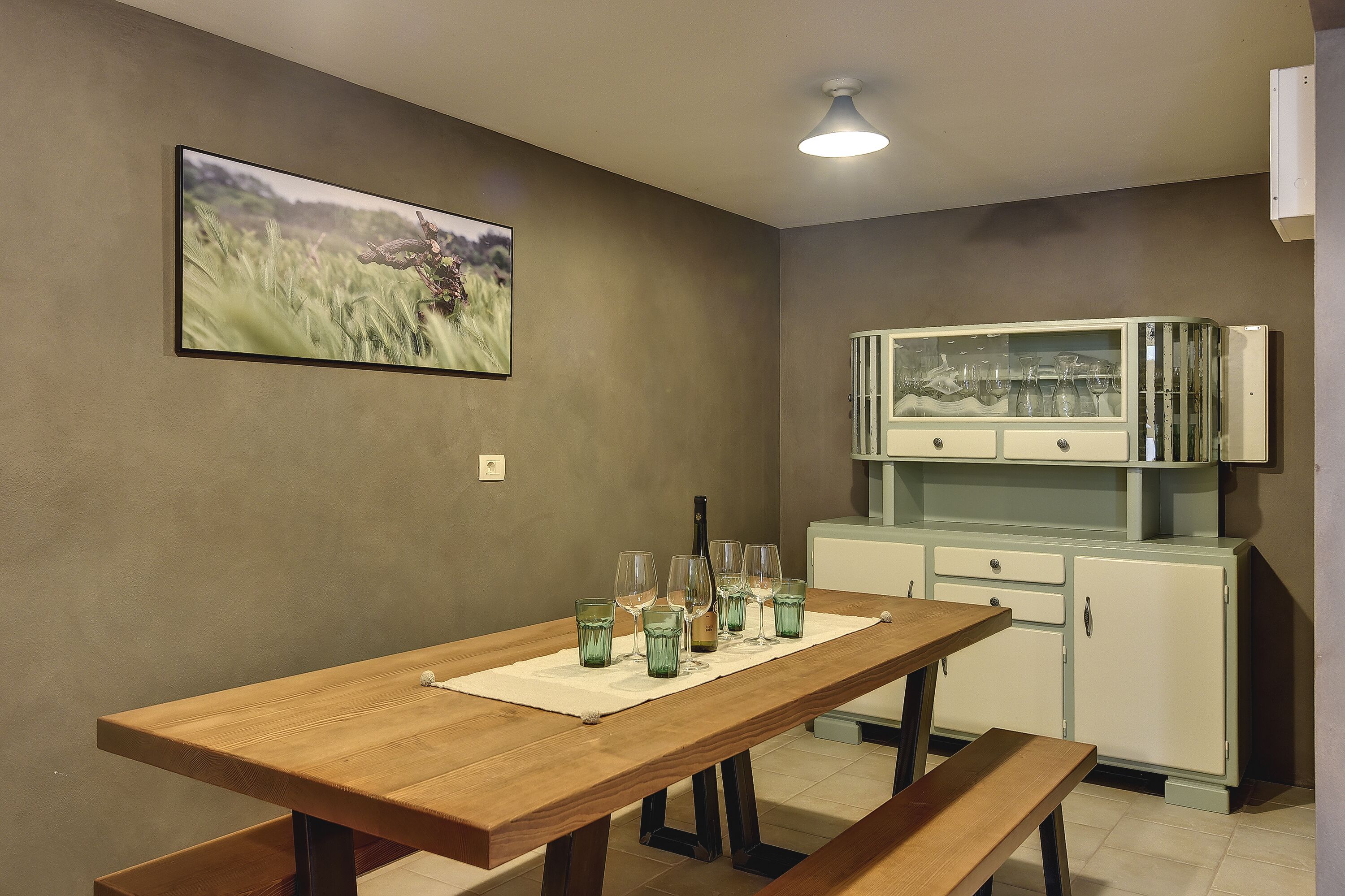 Uma sala de jantar com mesa de madeira, bancos e um armário com copos.