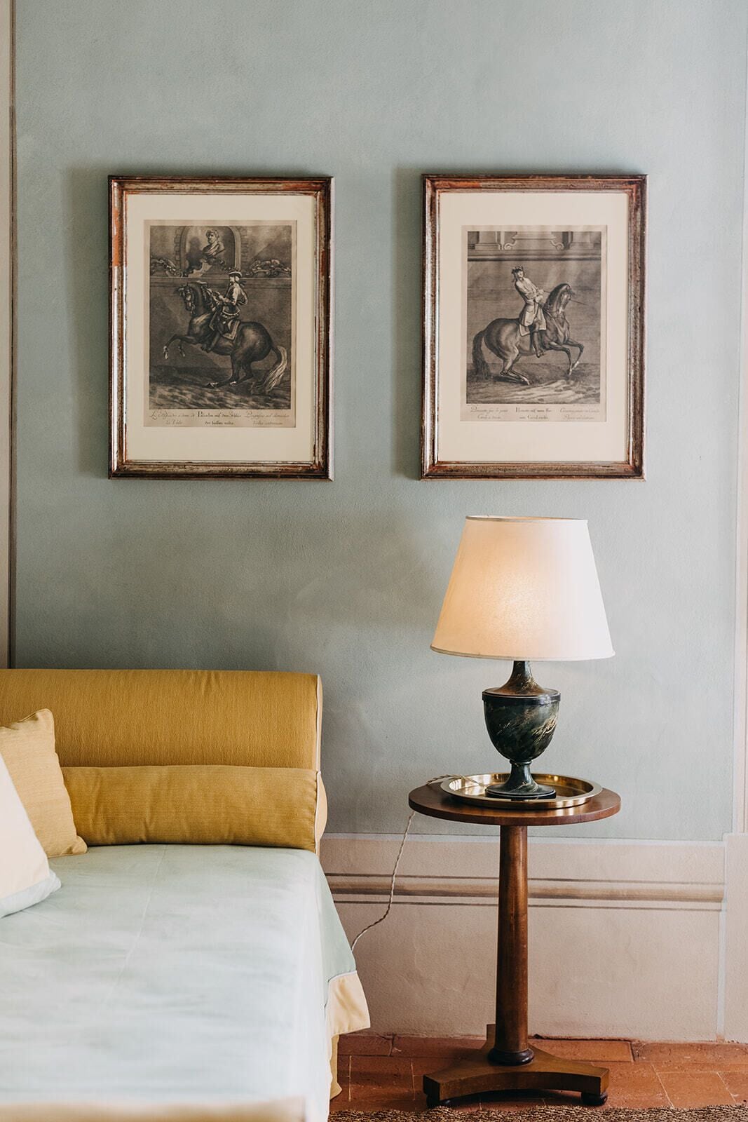 Una habitación con una cama, una mesa redonda con una lámpara y dos cuadros en la pared.