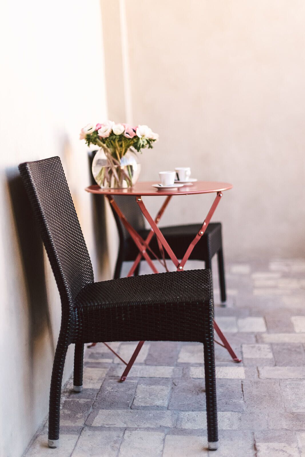 Una silla de mimbre negra y una mesita redonda con un florero y dos tazas blancas.