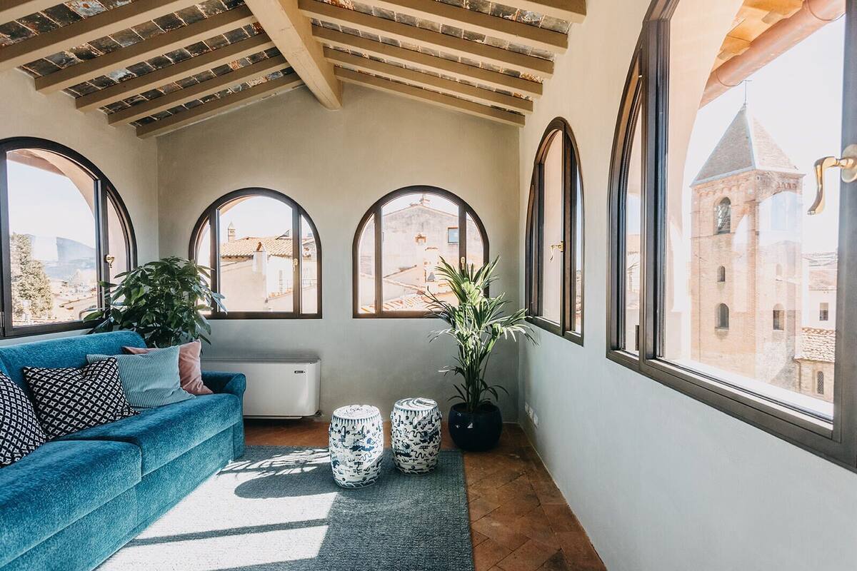 A cozy room with a blue sofa, two potted plants, and a view of historic buildings through large arched windows.
