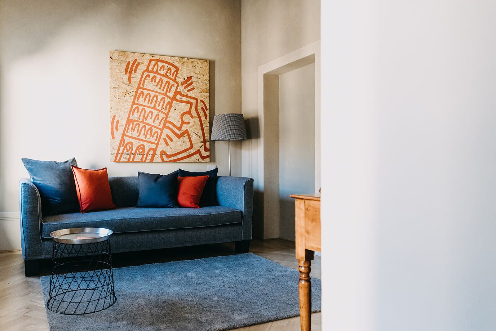 A living room with a blue sofa, red and blue cushions, a metal tray, a wooden side table, and a wall art piece.