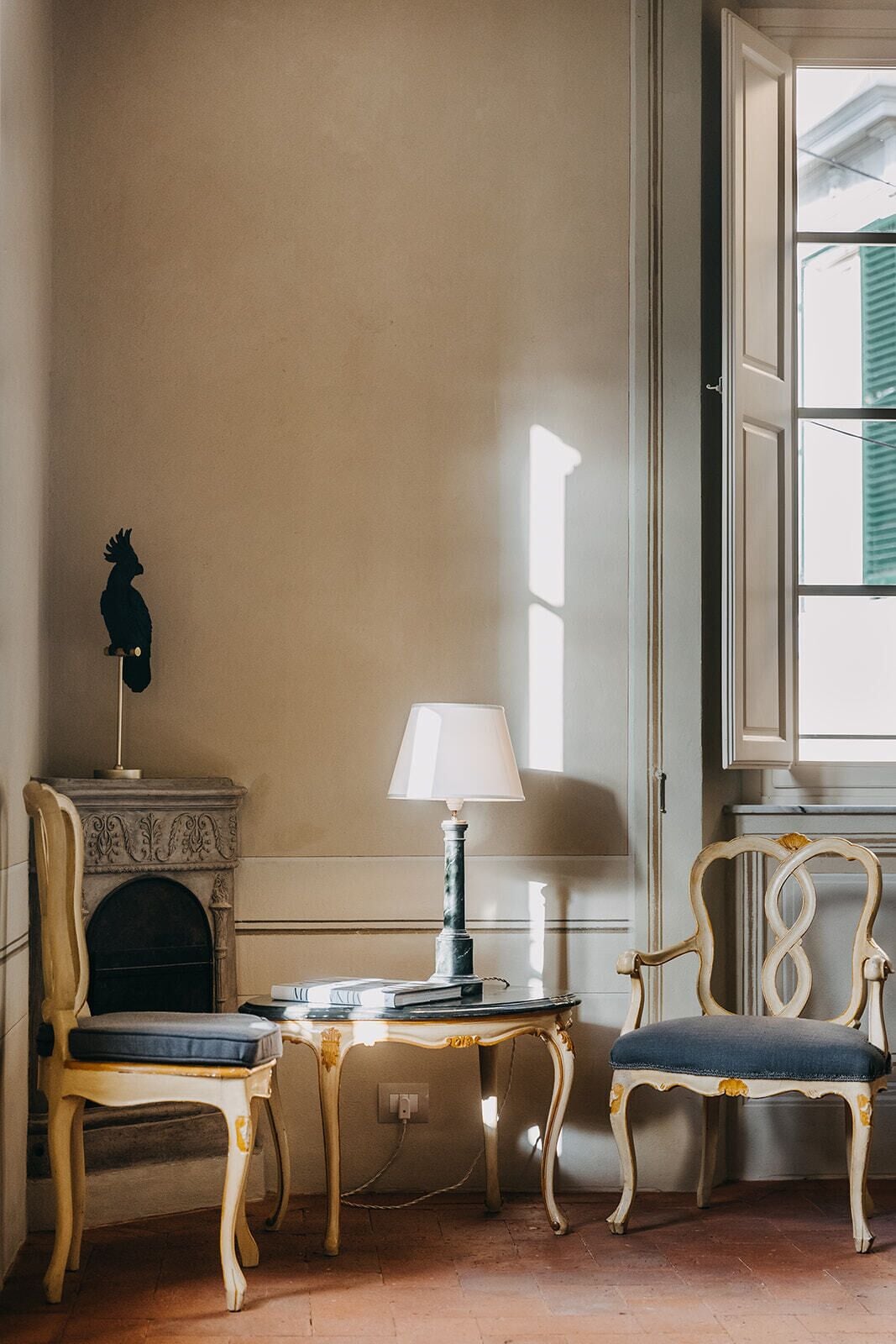 Une pièce avec une cheminée, une table avec une lampe, deux chaises et une fenêtre avec des stores.