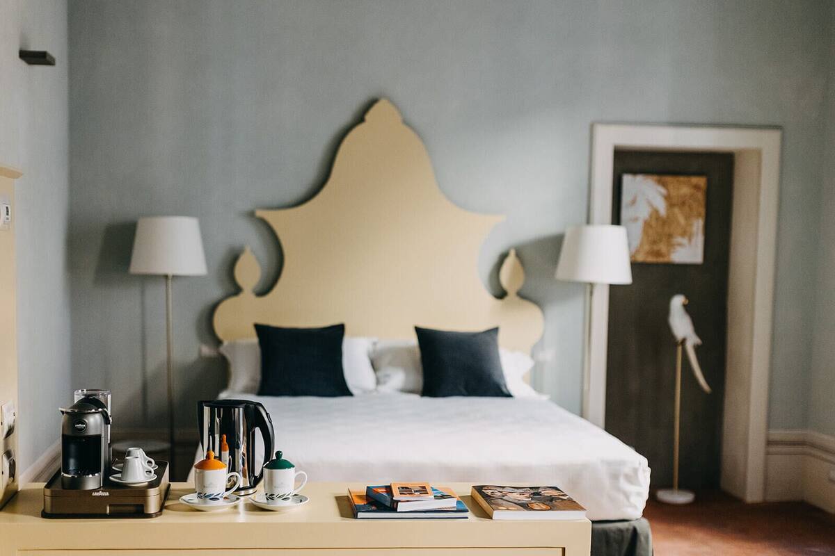 A neatly made bed with a decorative headboard, a bedside table with a coffee maker, cups, and books, and a lamp on either side of the bed.