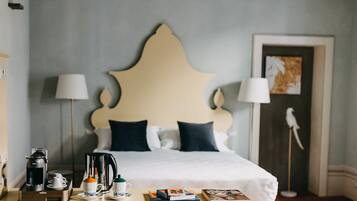 Una cama bien hecha con un cabecero decorativo, una mesita de noche con cafetera, tazas y libros, y una lámpara a cada lado de la cama.