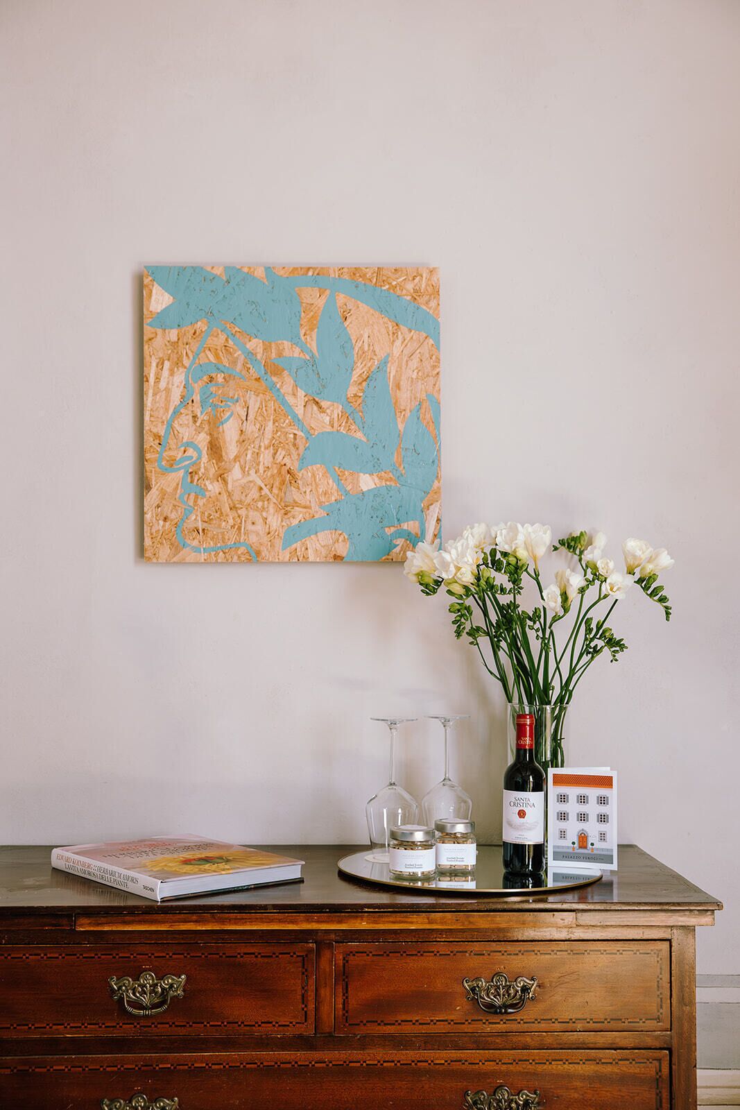 A wooden dresser with a book, wine glasses, a bottle, and a vase with flowers.