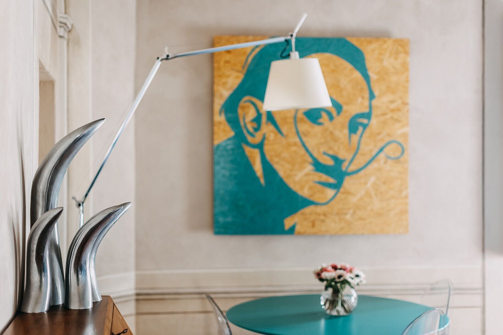 Une pièce avec une table de couleur turquoise, une lampe blanche, une œuvre d’art encadrée et un vase rempli de fleurs.