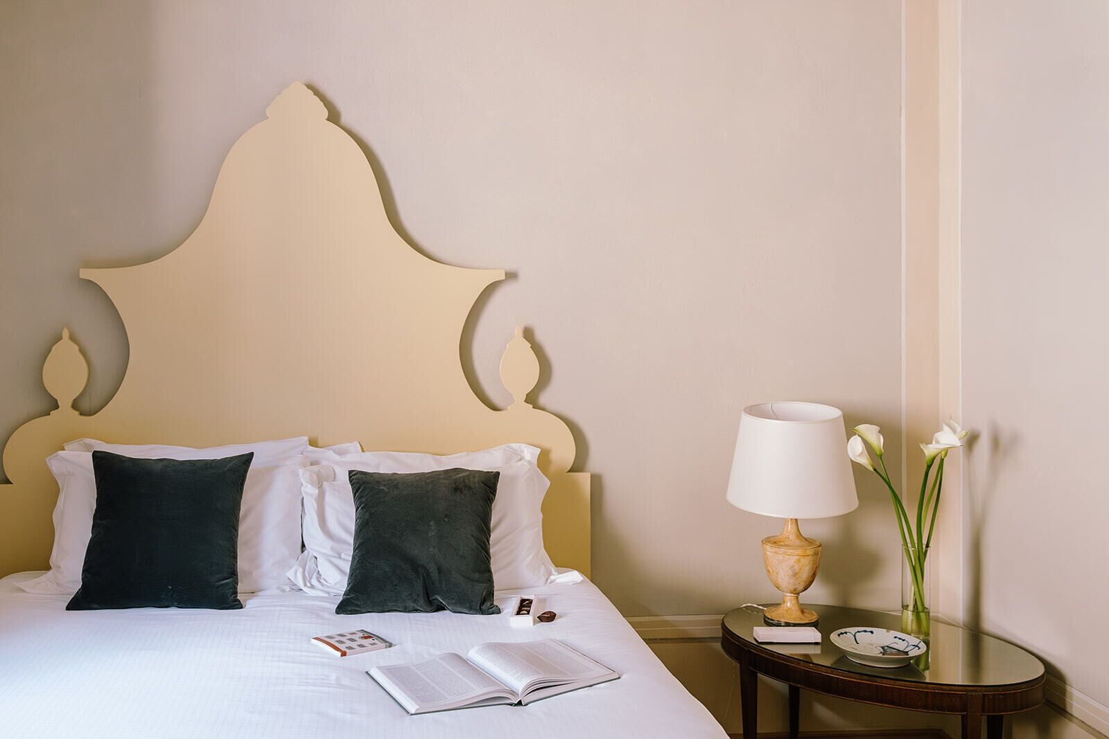 Una cama bien hecha con almohadas blancas y verde oscuro, una mesita de noche con una lámpara y un jarrón de flores, y un libro sobre la cama.