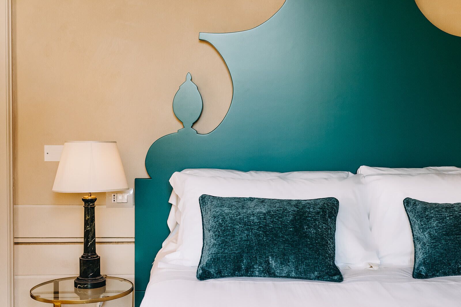 A neatly made bed with white linens and teal pillows, a bedside lamp, and a teal accent wall.