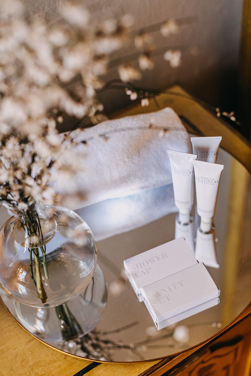 Un tocador de baño con una toalla blanca, un florero de cristal con flores y artículos de aseo.